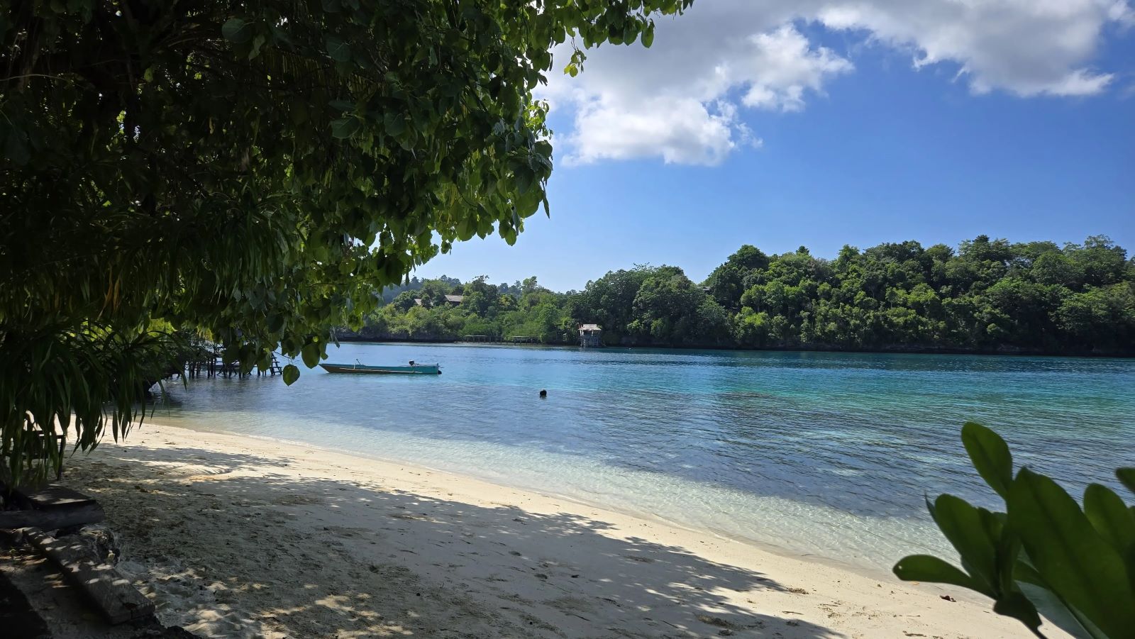 A shaded white-sand beach on a calm turquoise bay, with a small boat and forested islands across the water