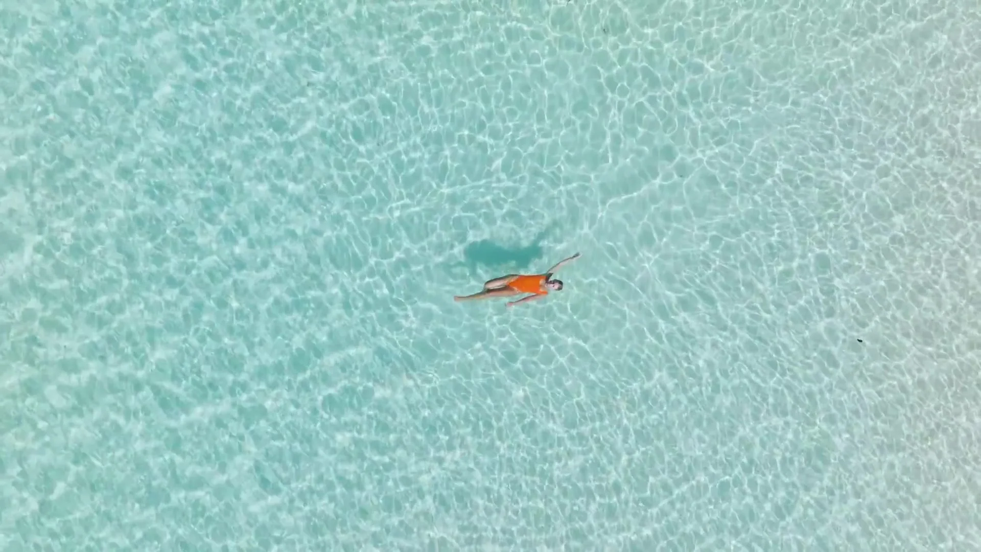 Aerial view of a swimmer in an orange swimsuit floating on her back in clear turquoise shallows
