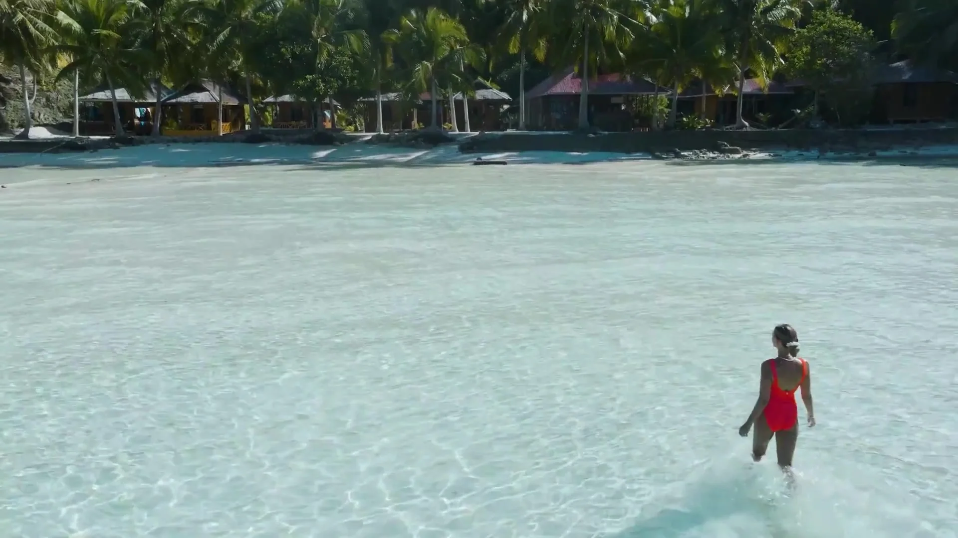 Woman in a red swimsuit wading through clear shallow water toward palm-shaded beach bungalows