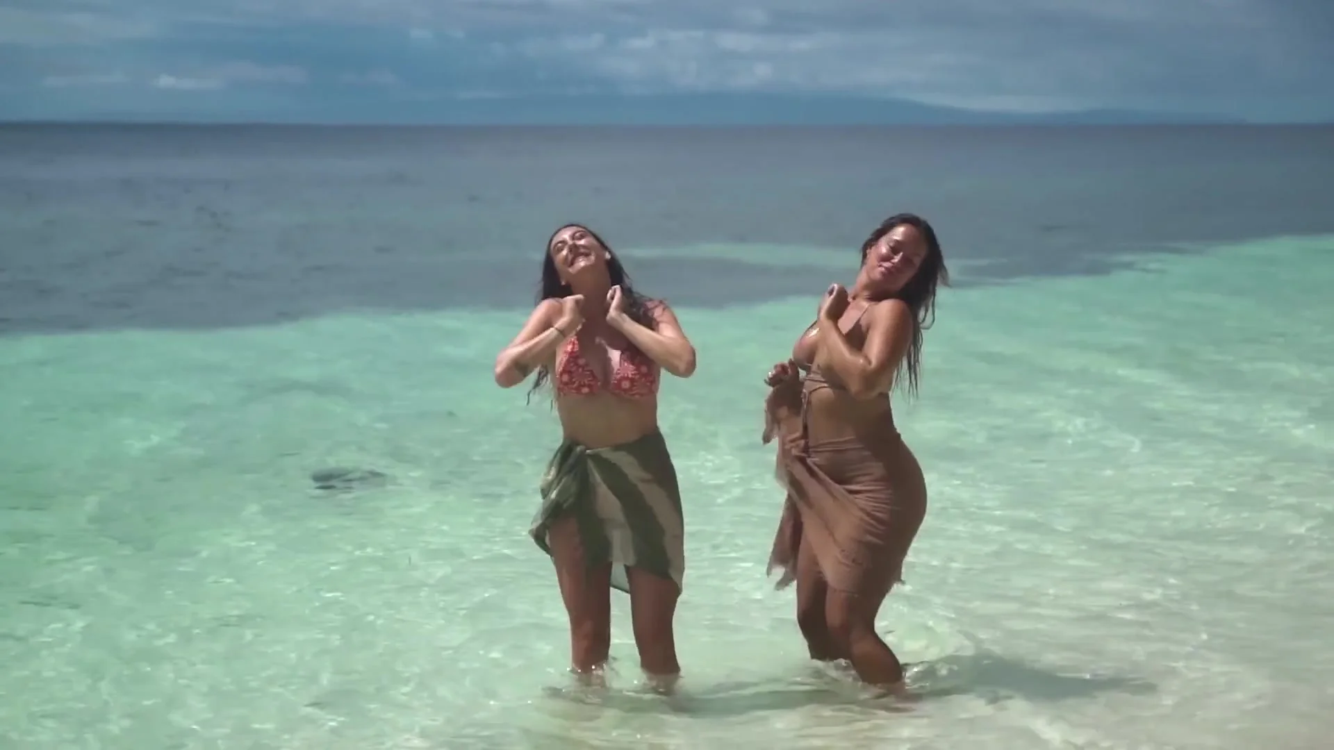 Two women in sarongs laughing and dancing knee-deep in clear turquoise water under cloudy skies
