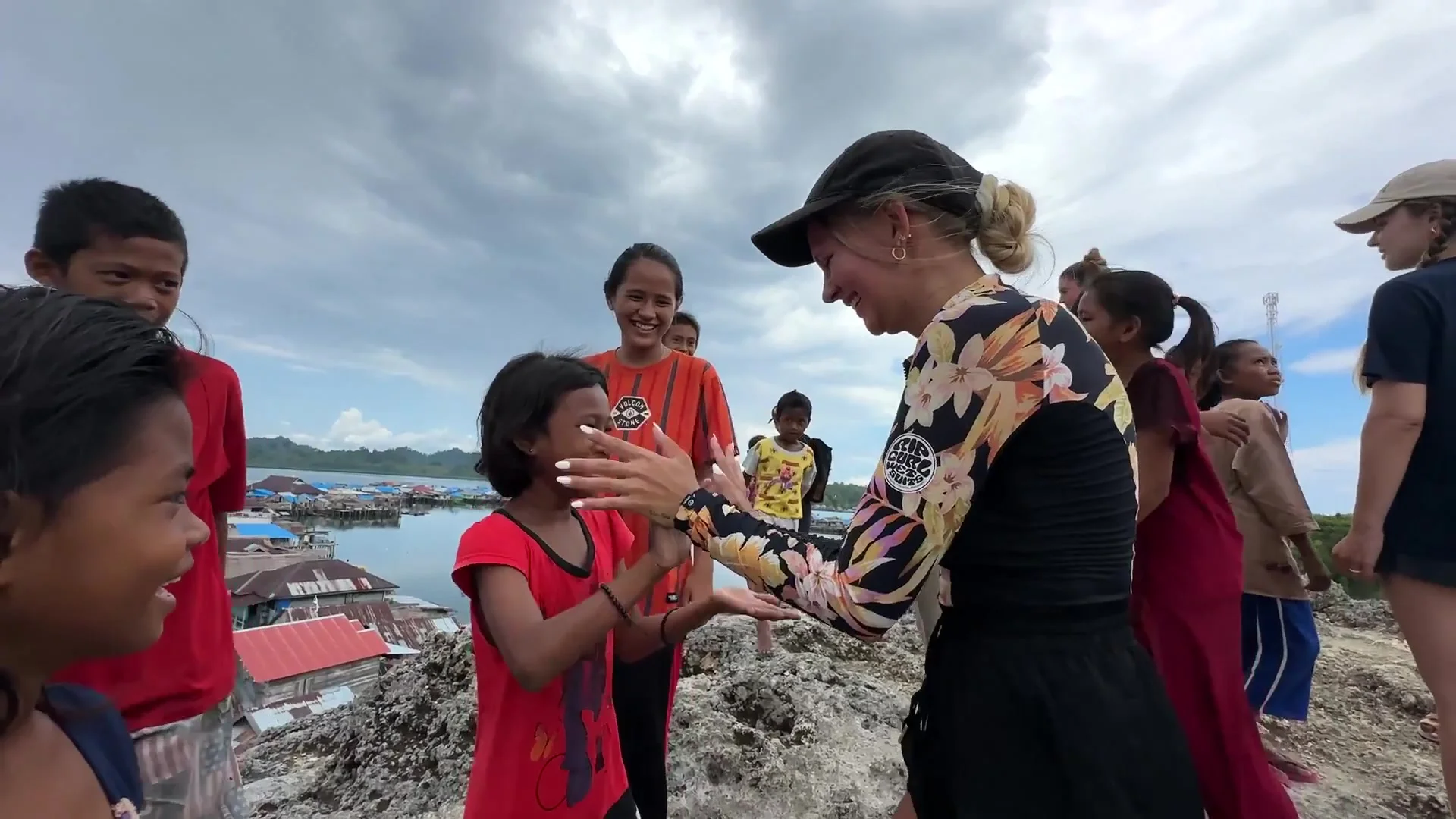 Traveller playing a clapping game with laughing children on a rocky hilltop above a stilt-house village