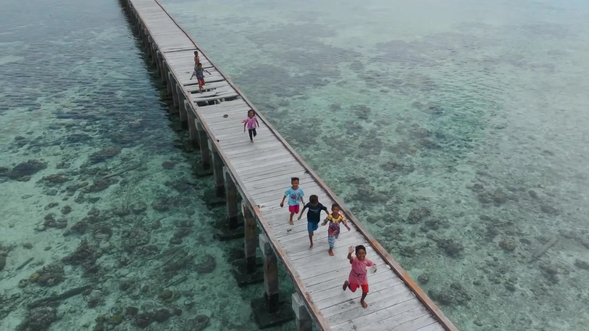 Aerial view of children running and waving along a long wooden jetty over clear reef shallows