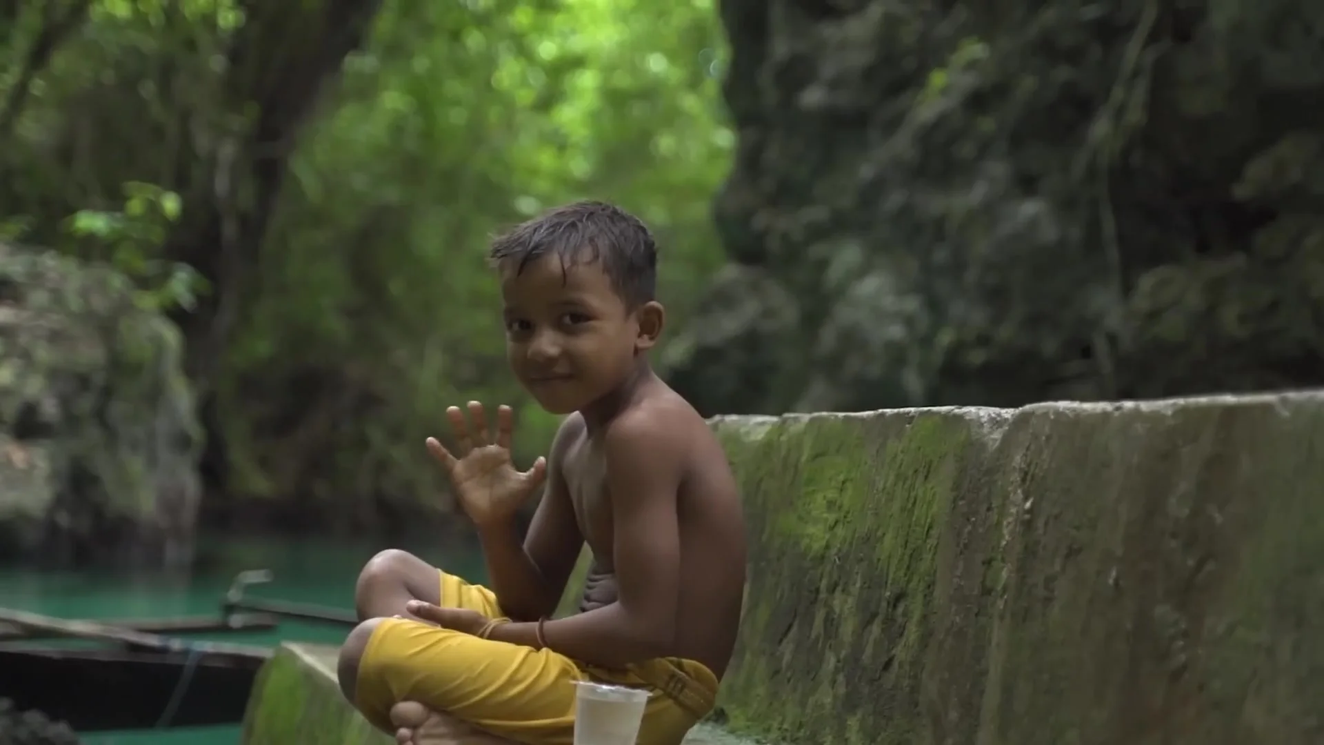 Smiling boy waving while sitting cross-legged on a mossy concrete wall beside green water