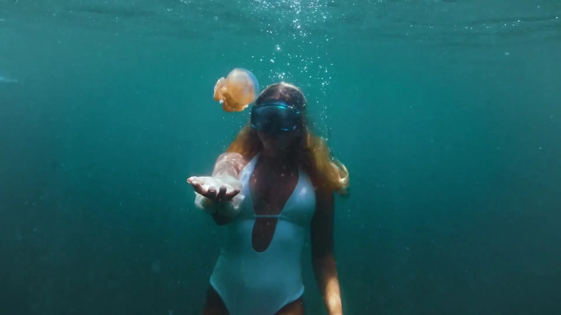 Snorkeller in a mask and white swimsuit reaching out a hand beside a drifting orange jellyfish
