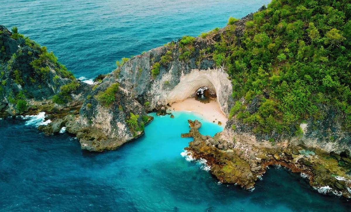 Hidden sandy cove tucked under a pale rock arch, ringed by turquoise water and green cliffs