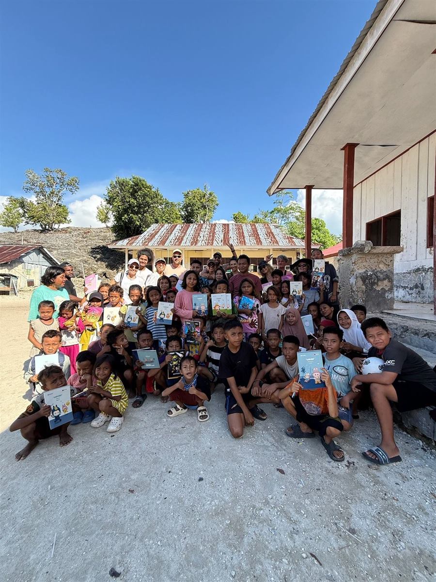 Travellers and village children holding up booklets outside a village building under a blue sky
