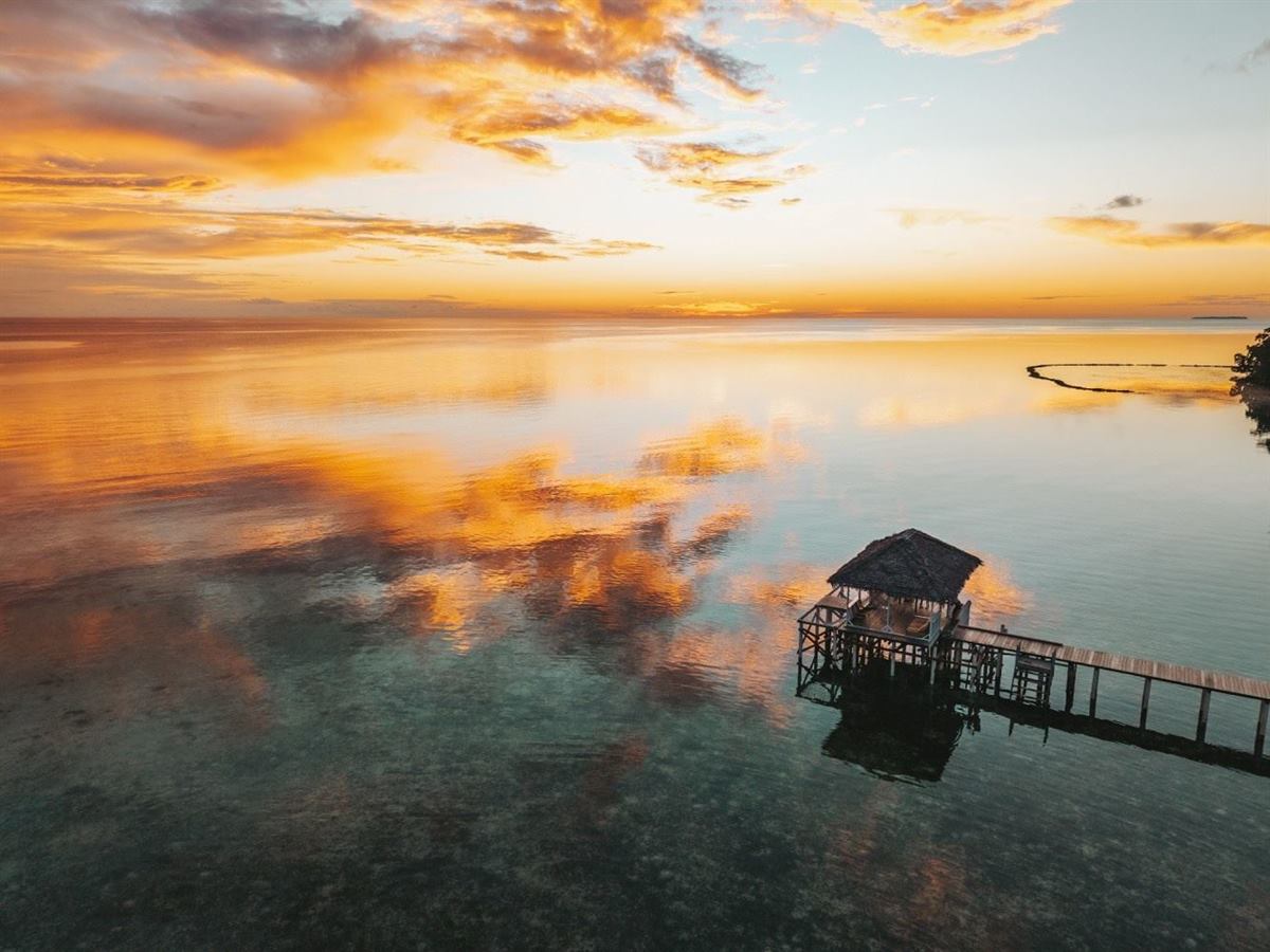 Thatched hut at the end of a wooden jetty over calm, glassy water at golden sunset