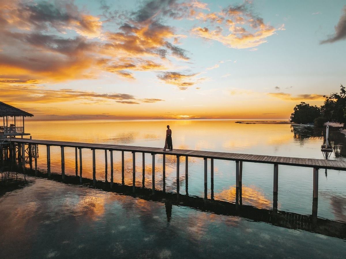 Person walking along a long wooden jetty over still water reflecting an orange sunset sky