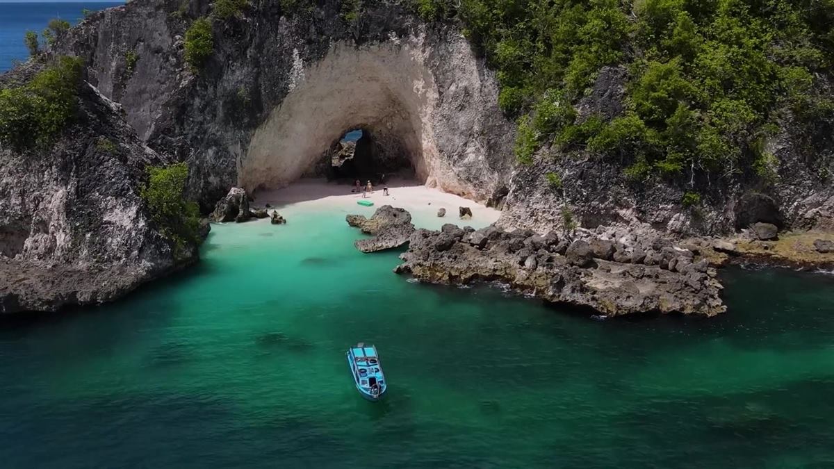 Aerial view of a speedboat anchored in a green cove below a small beach inside a rock arch