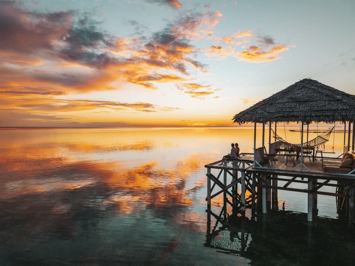 Two people sitting at the edge of a thatched over-water deck with a hammock at golden sunset
