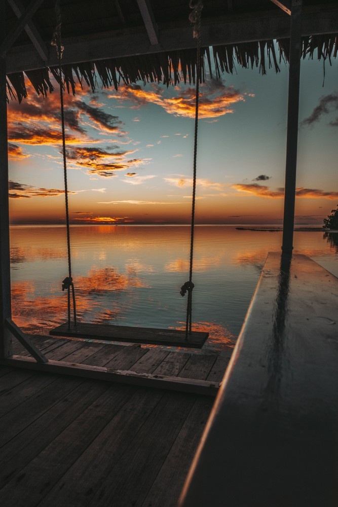 Rope swing hanging under a thatched roof, framing still water that reflects a fiery sunset